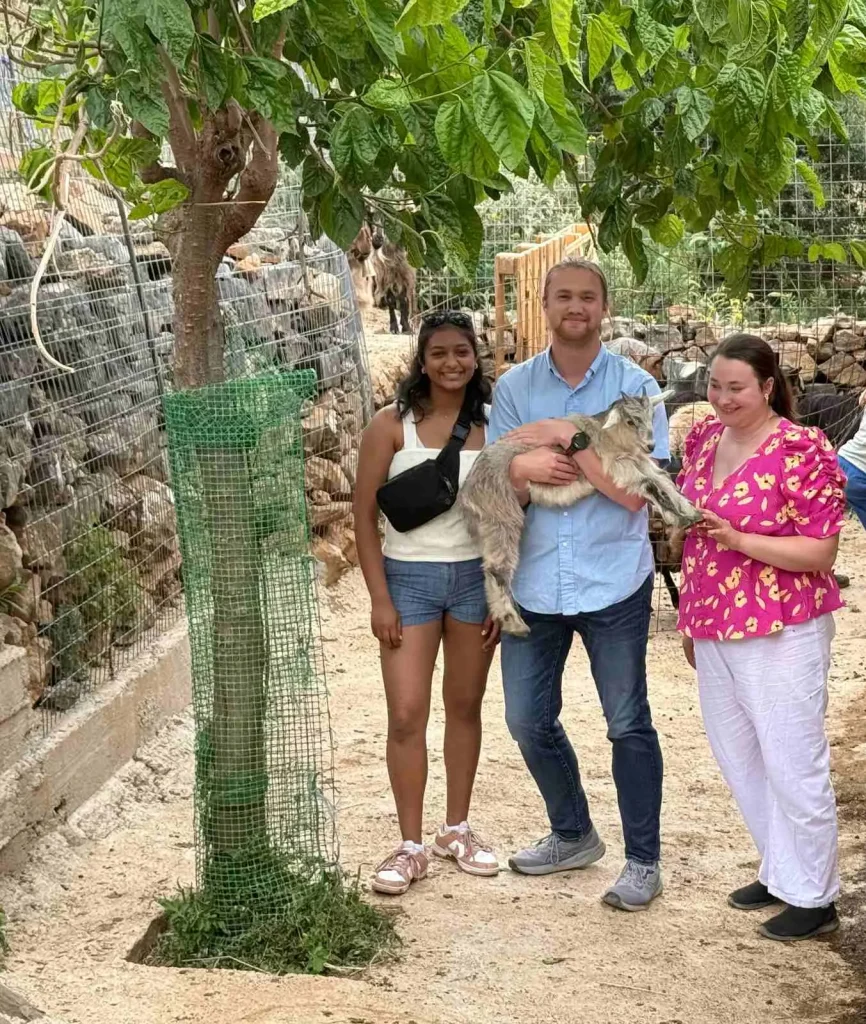 Three smiling visitors standing under a tree, with one holding a young goat during an interactive farm experience at Arevitis Farm in Crete.