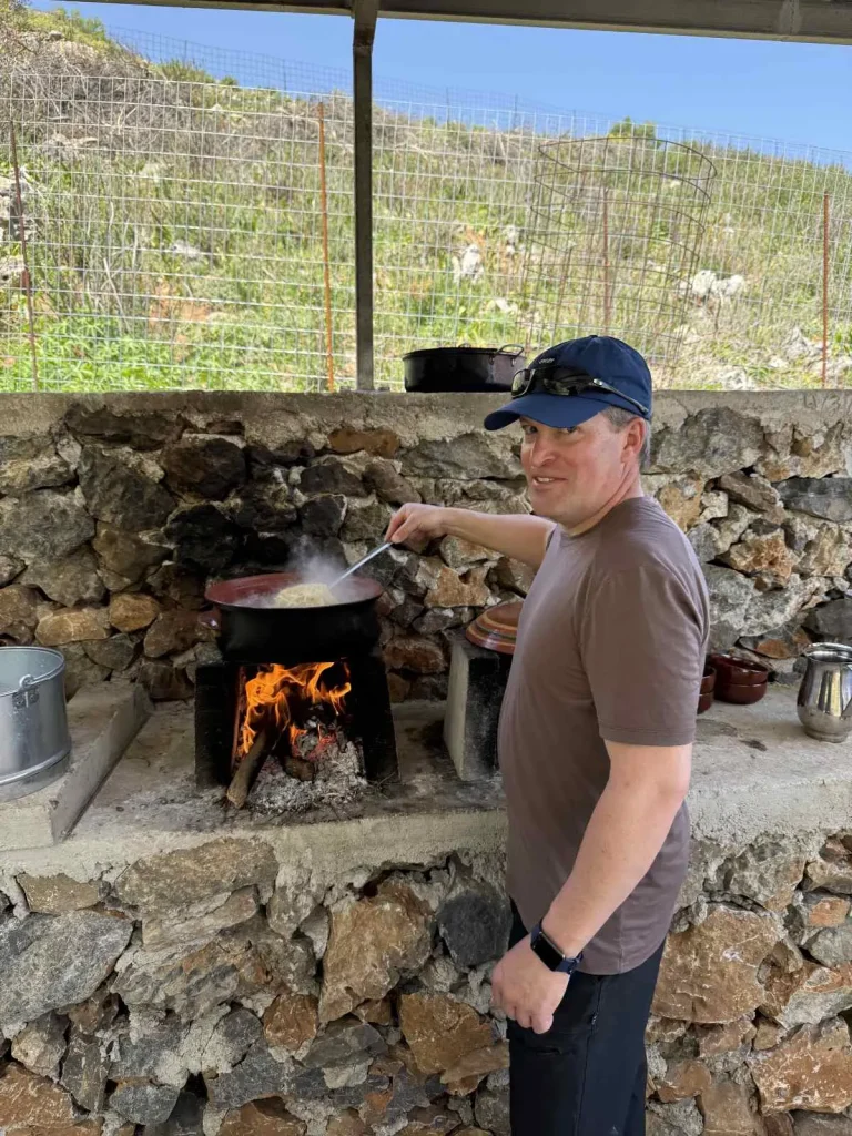 A visitor stirring a traditional dish in a clay pot over an open flame at an outdoor stone cooking area in rural Crete.