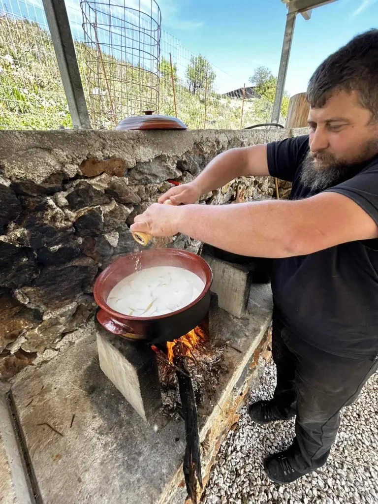 Man preparing traditional Cretan food in a clay pot over an open flame at an outdoor stone hearth at Arevitis Farm.
