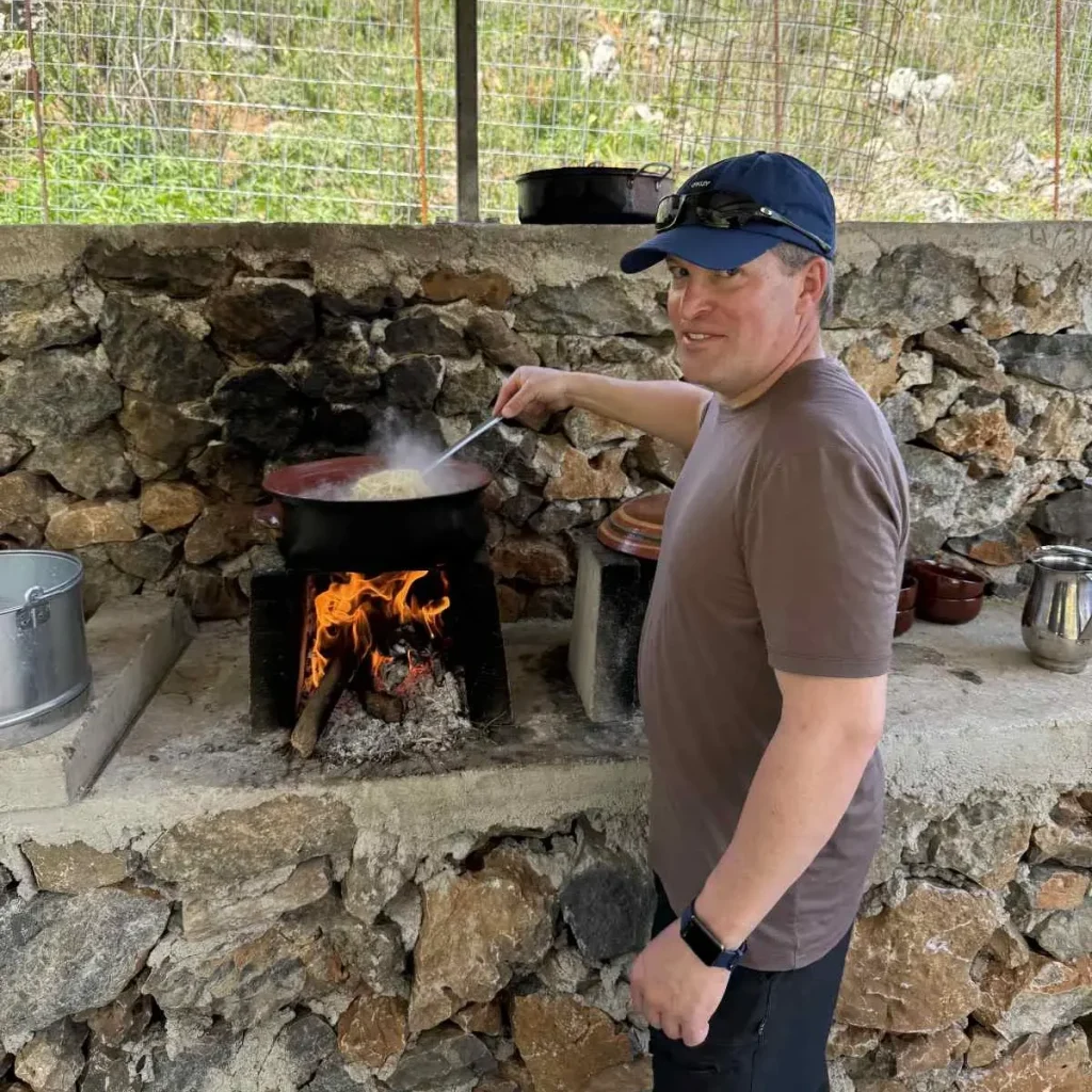 Ένας επισκέπτης ανακατεύει ένα παραδοσιακό φαγητό σε πήλινη κατσαρόλα πάνω σε ανοιχτή φωτιά, σε υπαίθρια πετρόκτιστη εστία στην αγροτική Κρήτη. Μαγειρική Εμπειρία